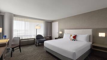 A hotel room with a large bed, a desk with a chair, a standing lamp, and a window with curtains.