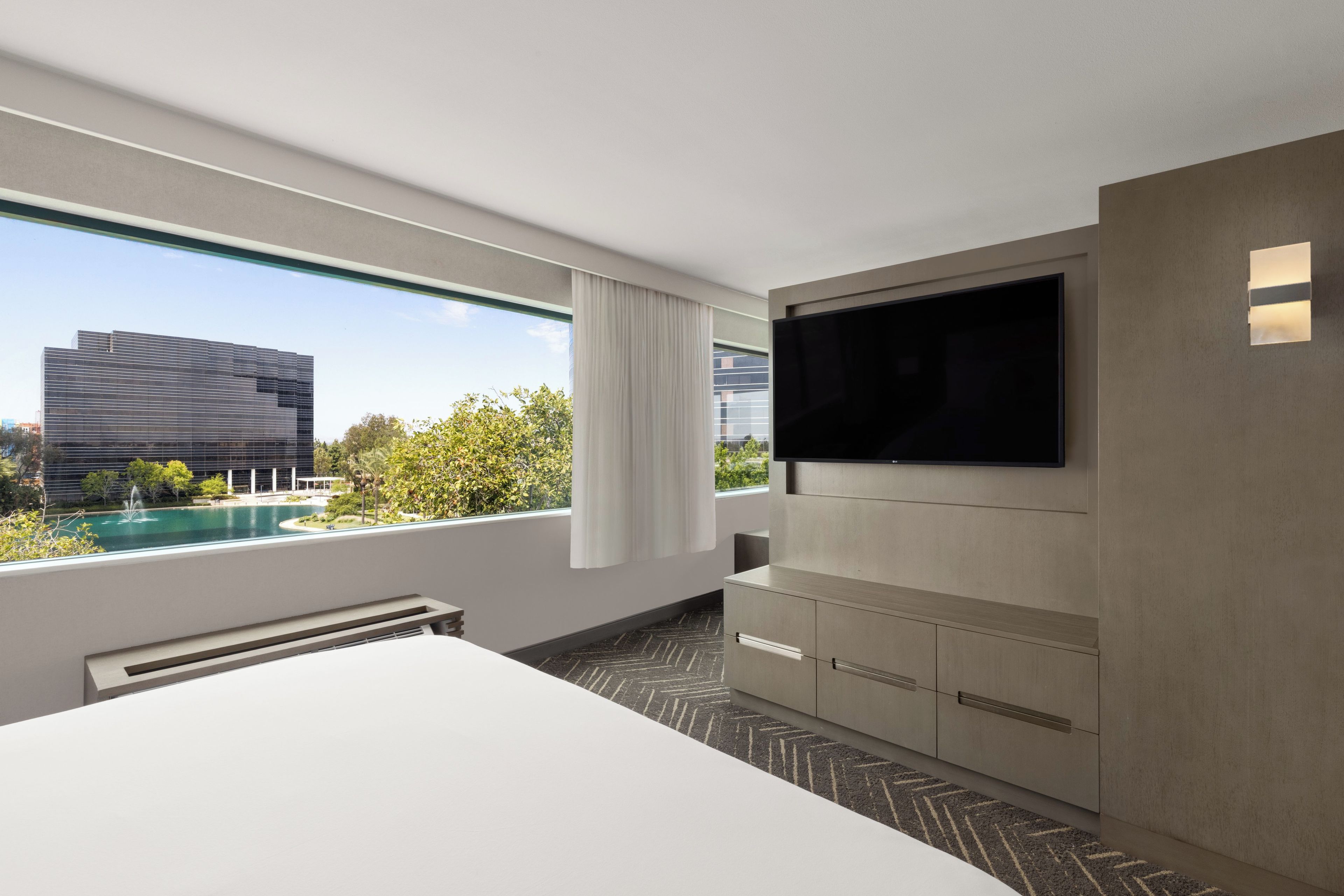 A modern hotel room with a large bed, a flat-screen TV mounted on the wall, and a view of a swimming pool and buildings.