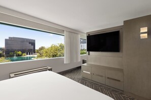 A modern hotel room with a large bed, a flat-screen TV mounted on the wall, and a view of a swimming pool and buildings.