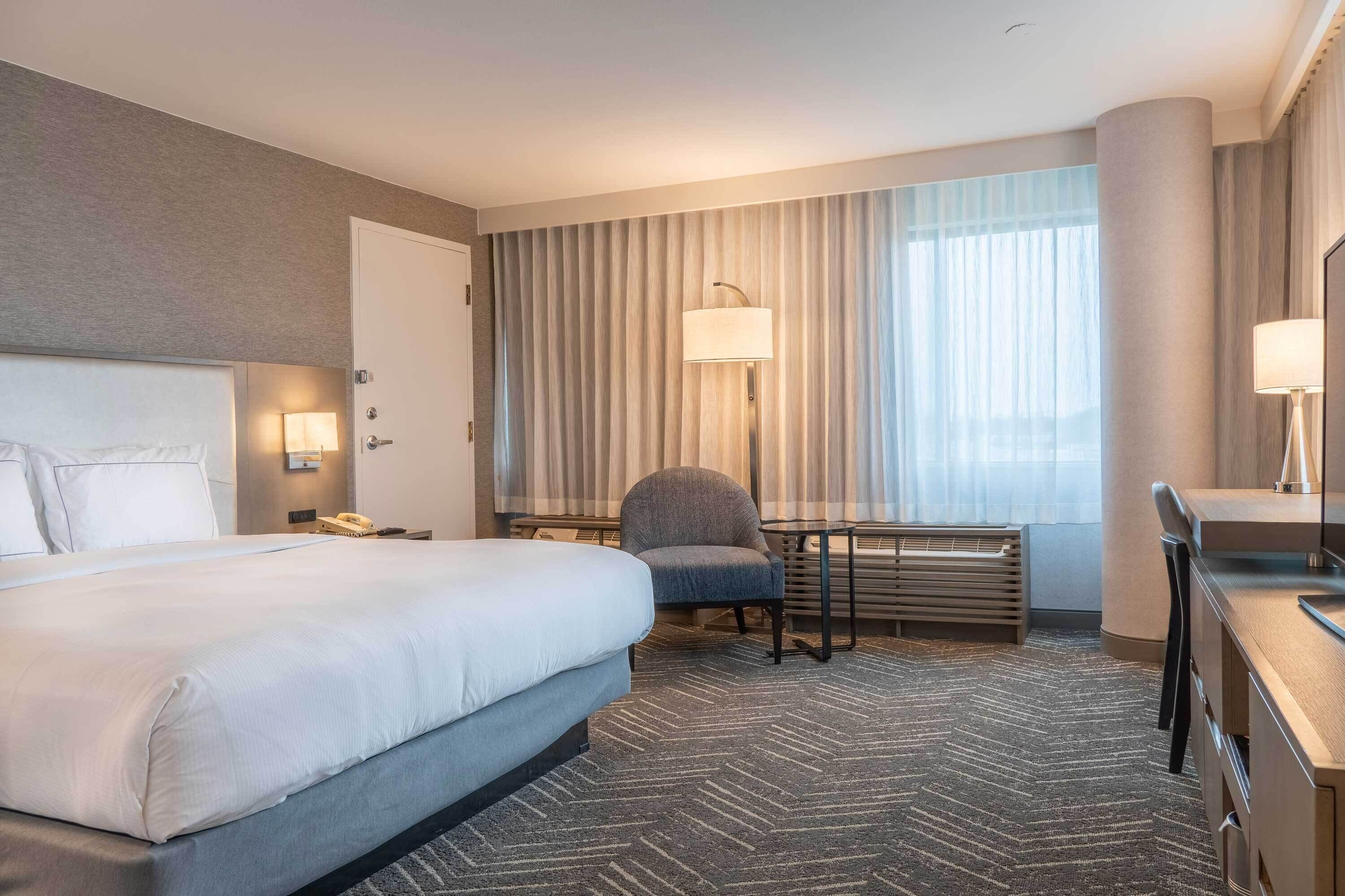 Habitación de hotel con una cama grande, un escritorio, una silla, una lámpara y una ventana con cortinas.