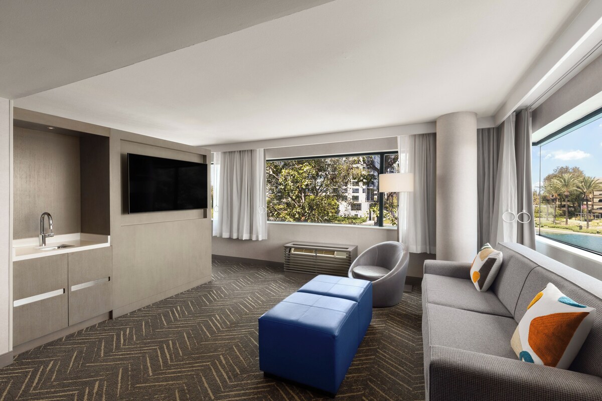 A hotel room with a kitchenette, a flat-screen TV, a sofa with cushions, a blue ottoman, a chair, and a large window with a view of buildings and trees.