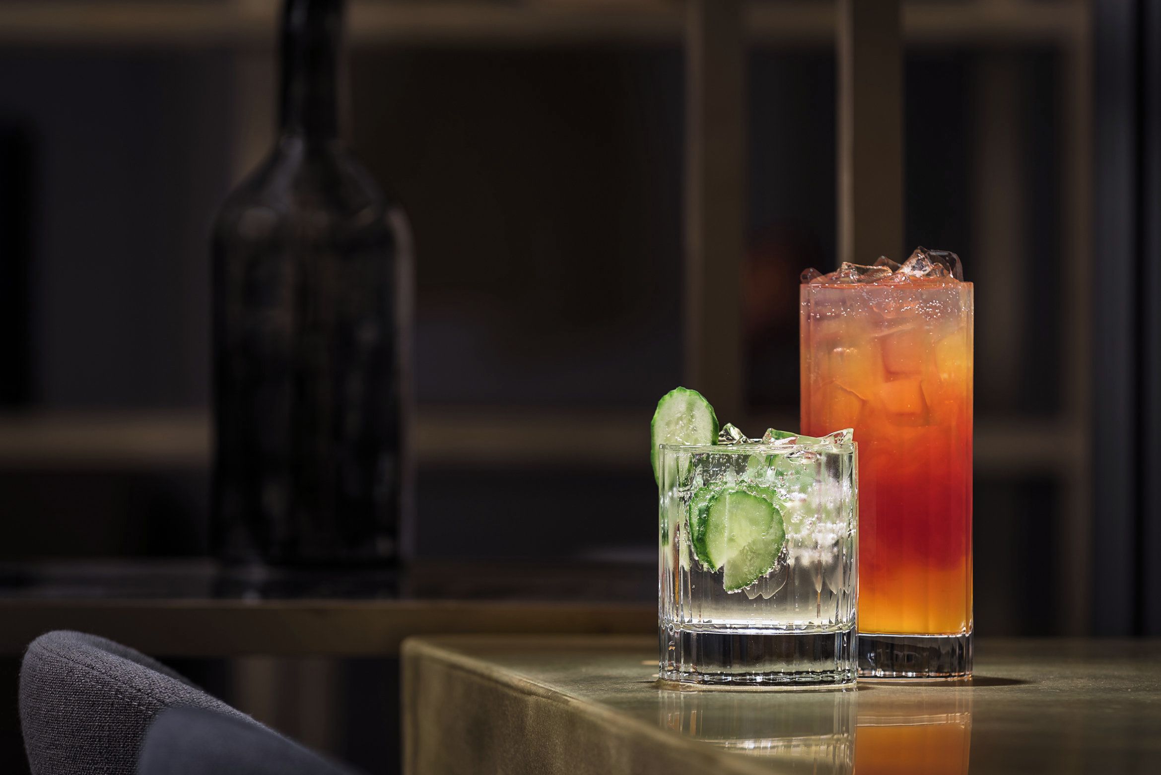 Two glasses of beverages on a table with a bottle in the background.