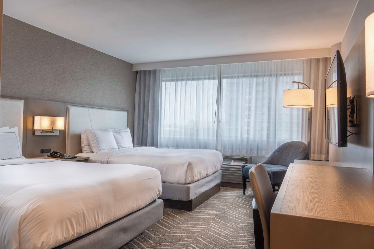 A hotel room with two beds, a desk, a chair, a TV, and a large window with curtains.