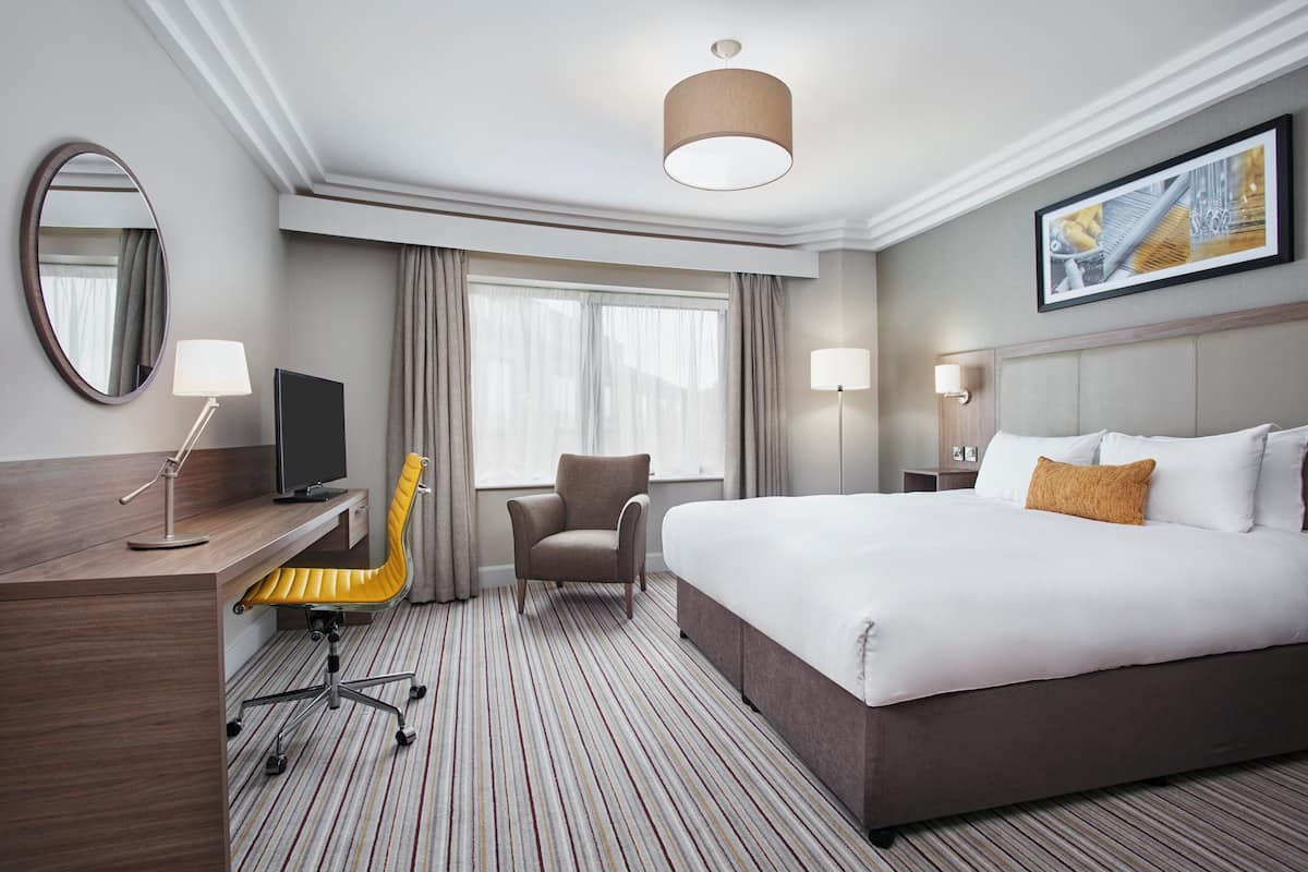 Ein Hotelzimmer mit einem großen Bett, einem Schreibtisch mit Stuhl, einem Fernseher und einem großen Fenster mit Vorhängen.