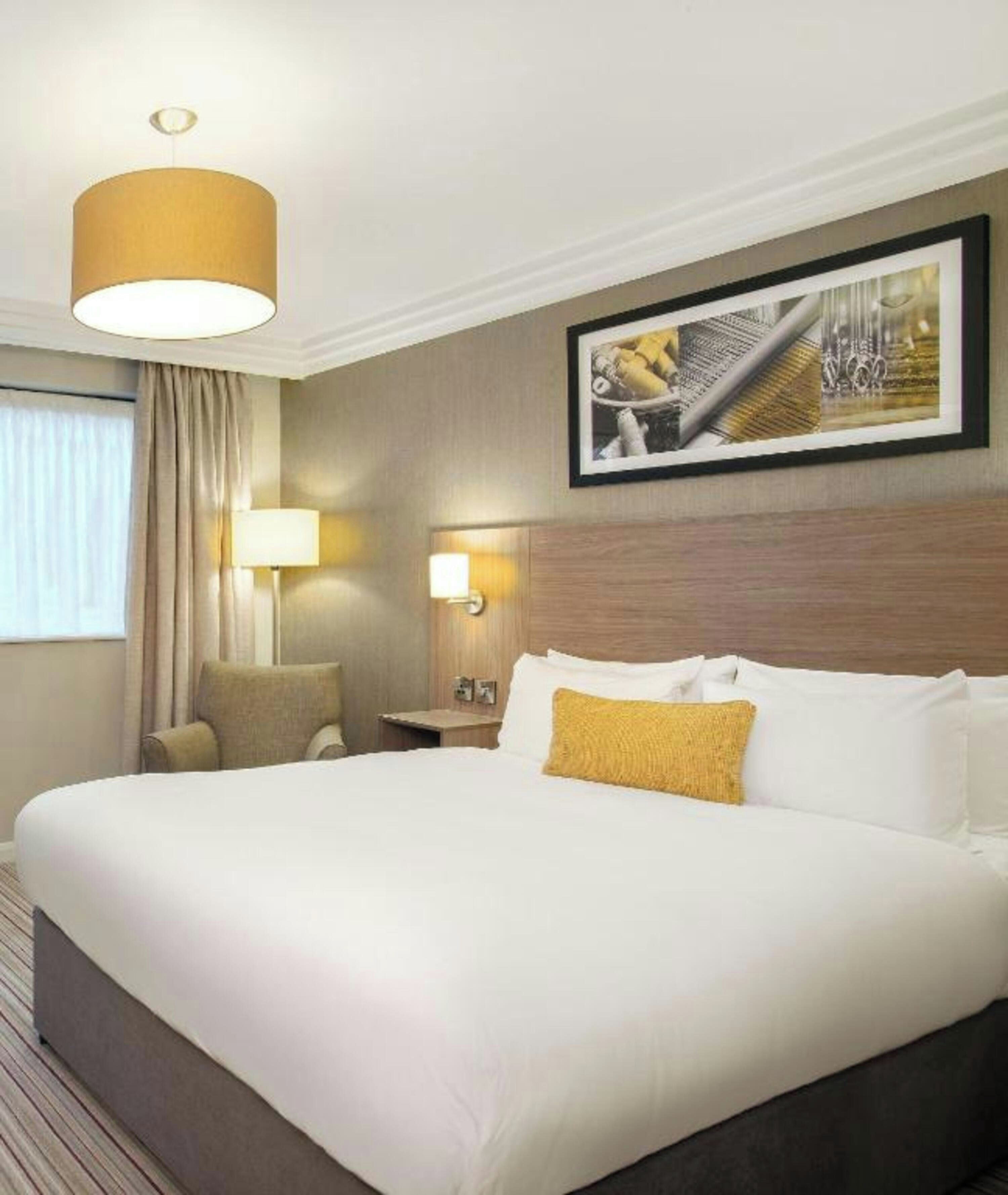 A hotel room with a large bed, a bedside lamp, a chair, a nightstand, and a framed picture on the wall.