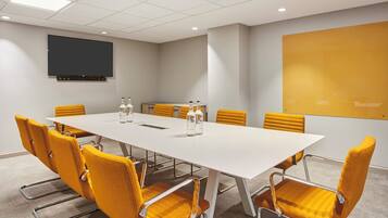 A conference room with a long table, six yellow chairs, a projector screen, and a flat-screen TV on the wall.