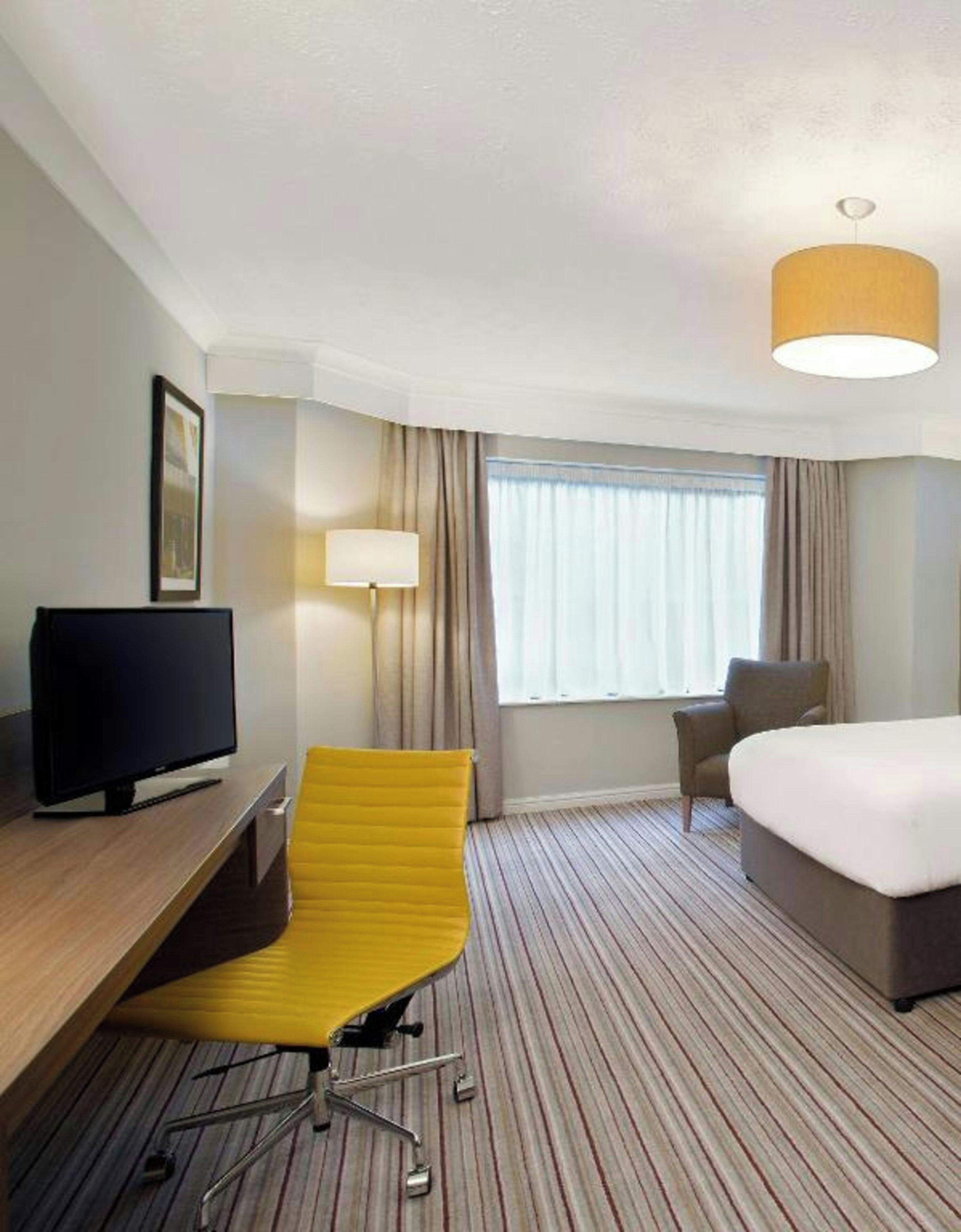 Habitación de hotel con cama, escritorio con televisor, una silla amarilla, lámpara y ventana con cortinas.