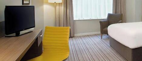 Habitación de hotel con cama, escritorio con televisor, una silla amarilla, lámpara y ventana con cortinas.