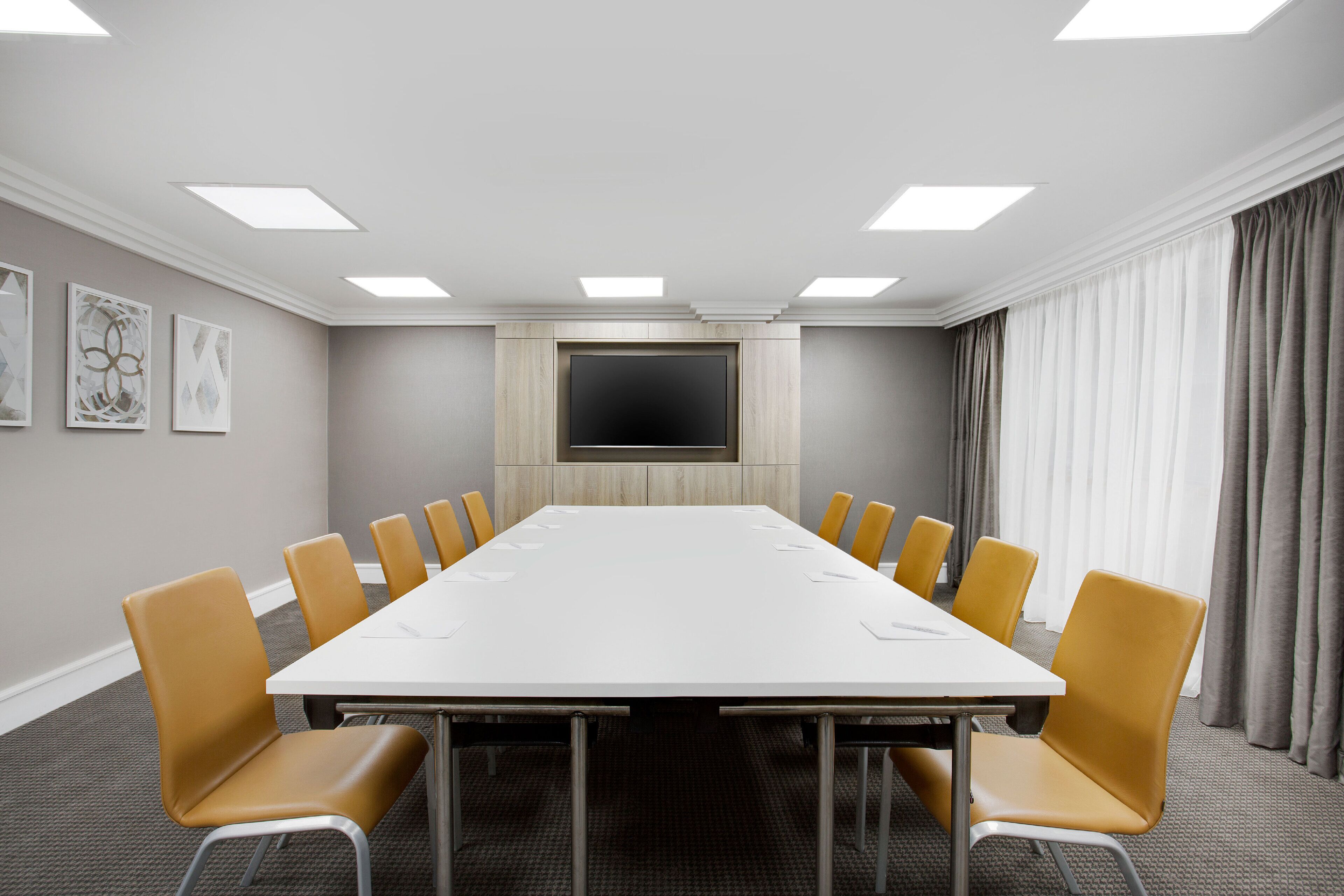 A conference room with a long table, yellow chairs, a flat-screen TV, and a large window with curtains.