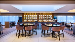 A modern bar area with round tables and chairs, a wine display, and a view of the city through large windows. - Leonardo Hotel and Conference Venue Hinckley Island (Hinckley)