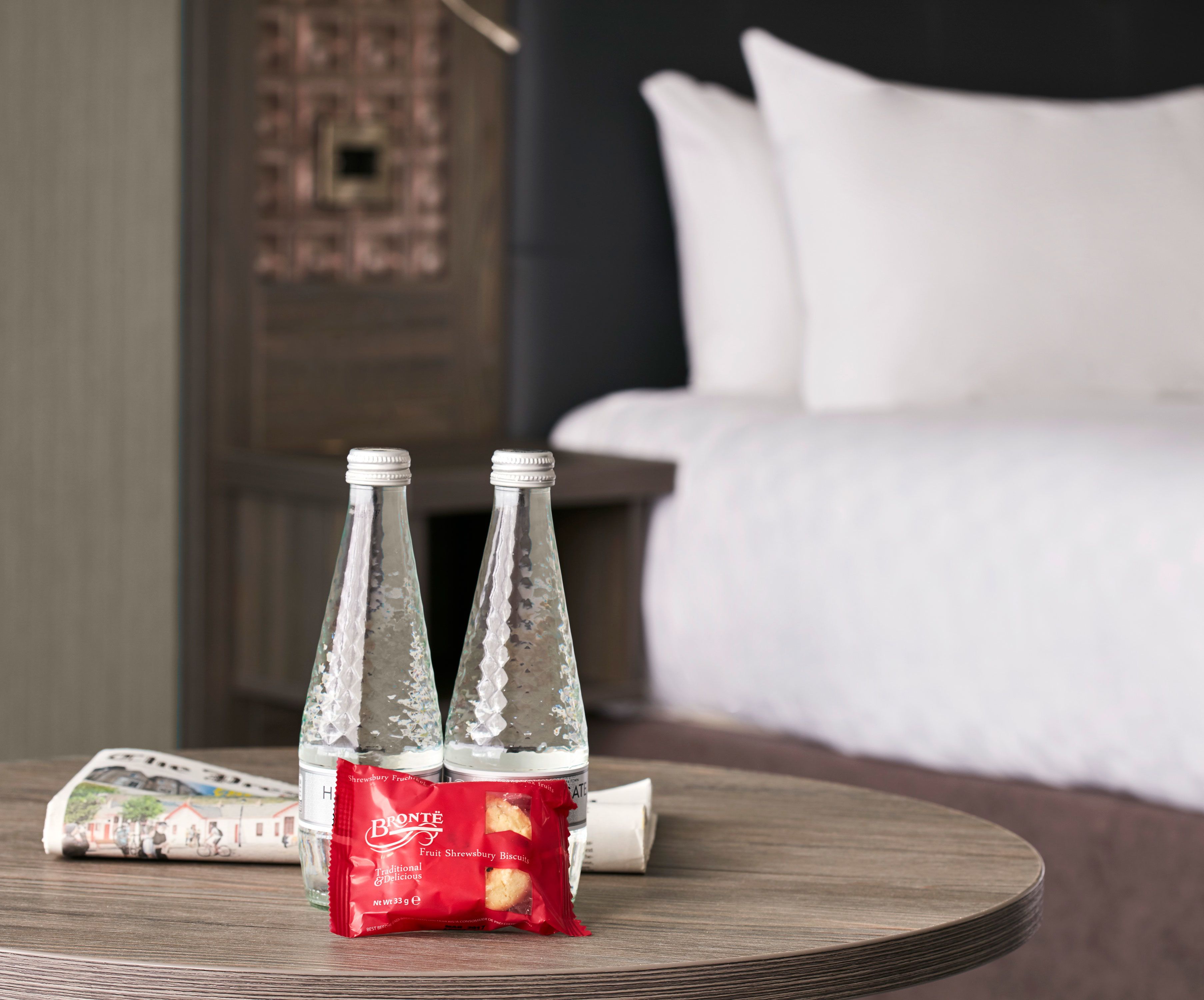Una habitación de hotel con una mesa redonda de madera, dos botellas de vidrio y una bolsita de galletas.