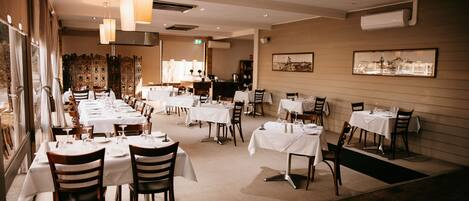 A restaurant with multiple tables and chairs, arranged in a spacious dining area with a high ceiling and pendant lighting.