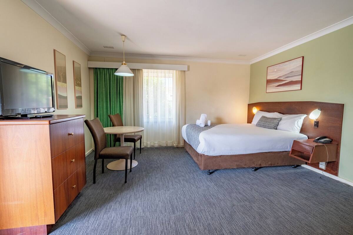 Ein Hotelzimmer mit einem Bett, einem Schreibtisch mit Fernseher, einem Sessel, einem kleinen Tisch und einem Fenster mit Vorhängen.