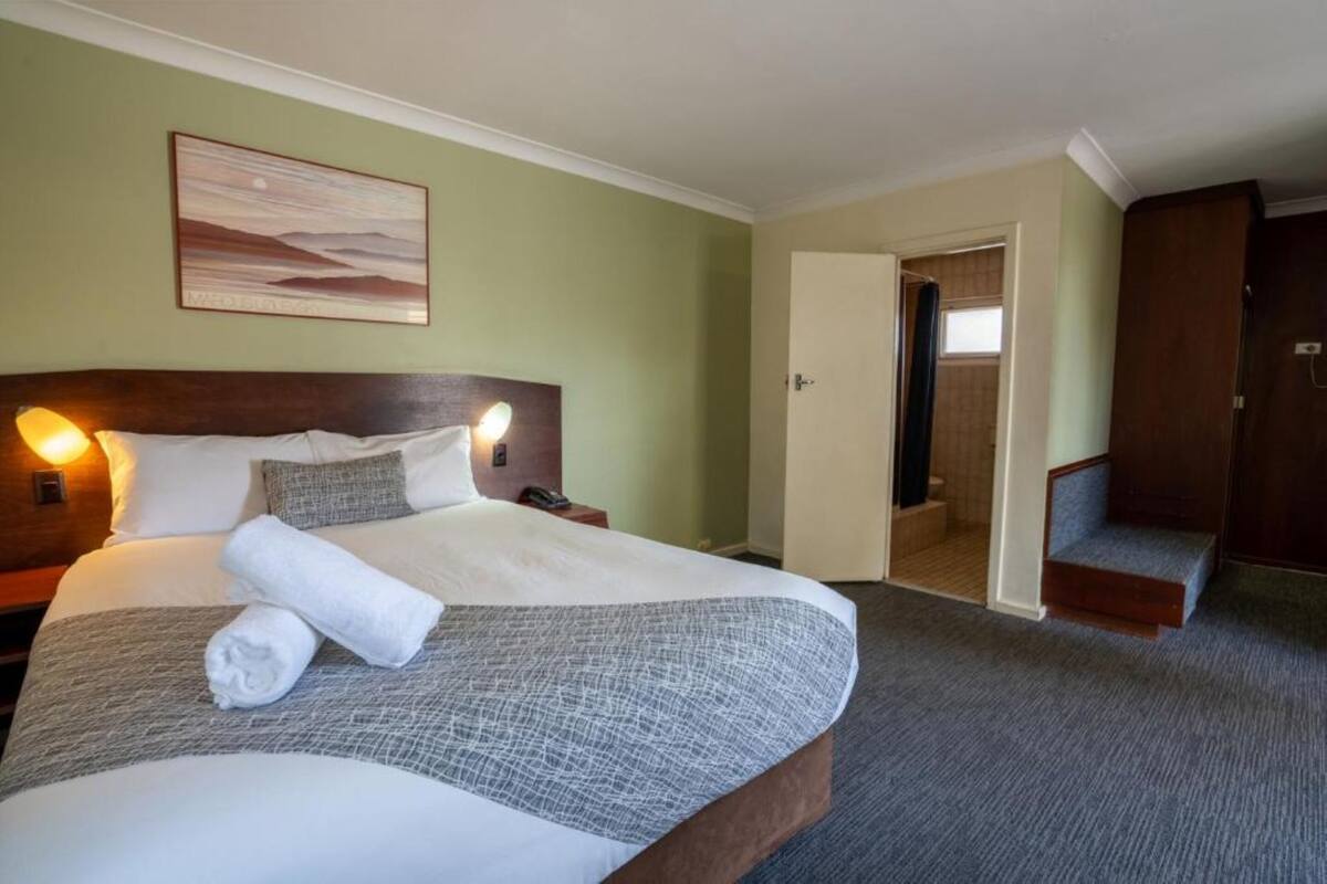 Ein Hotelzimmer mit einem großen Bett, einem Kopfteil, einem Nachttisch, einer Wandlampe, einem Gemälde und einer Tür zu einem Badezimmer.