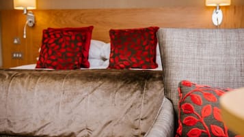 Una habitación de hotel con una cama, dos almohadas rojas con motivos de hojas, un sillón tapizado gris y lámparas fijadas a la pared.