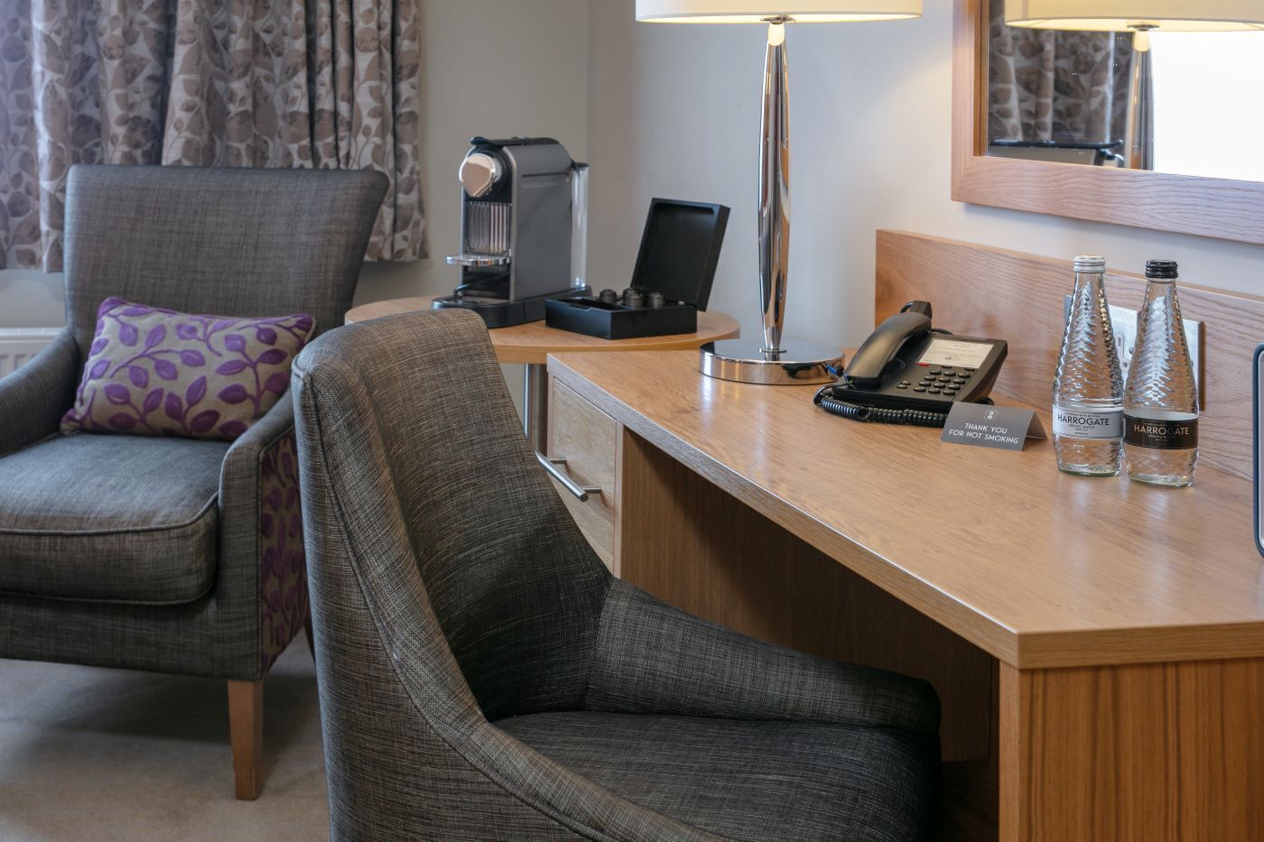 A hotel room with a wooden desk, two chairs, a lamp, a phone, and two Harrogate branded water bottles.
