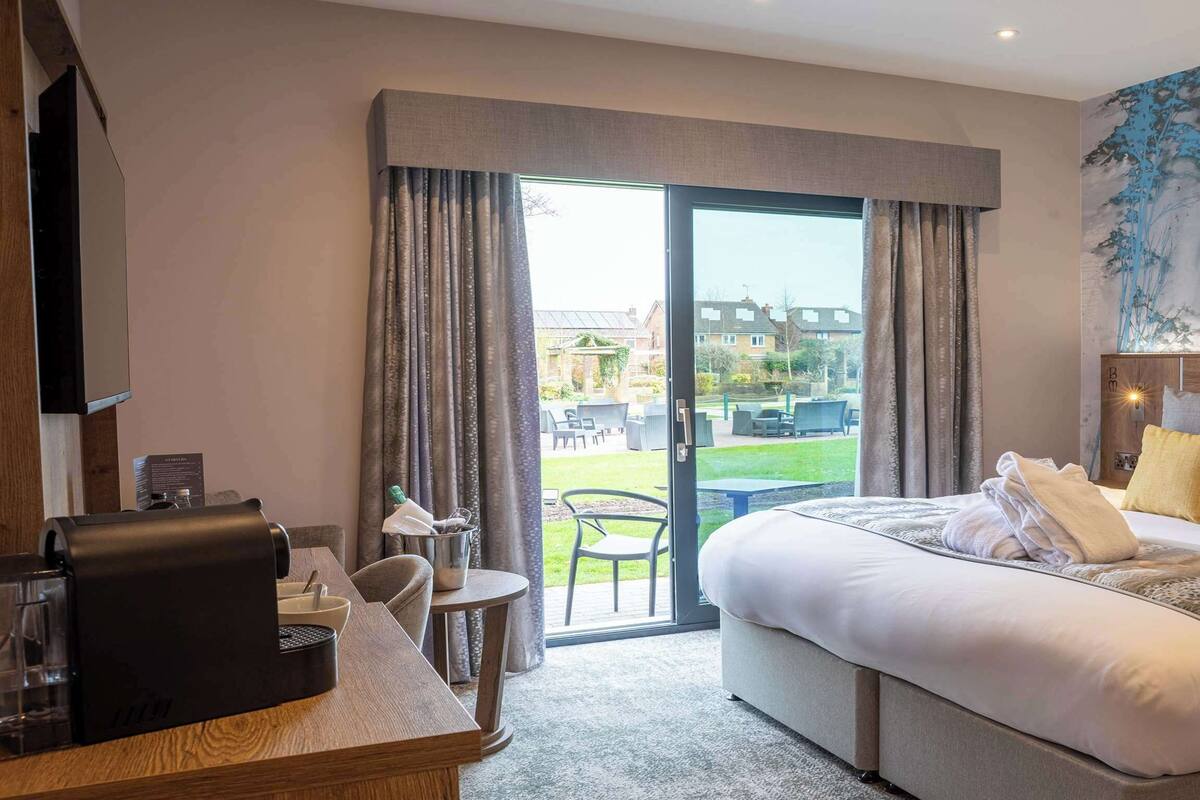A hotel room with a large bed, a desk with a coffee machine, a view of the outdoors, and a painting on the wall.