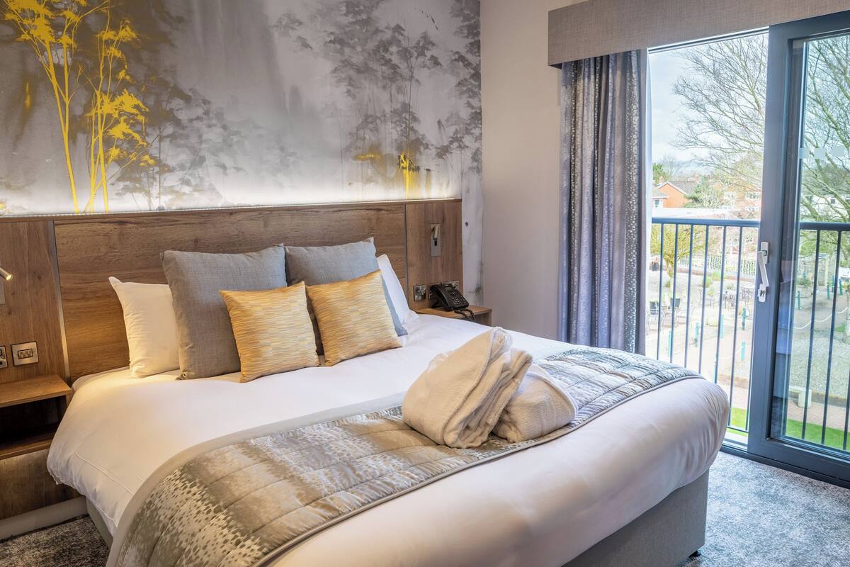 A hotel room with a large bed, a wooden headboard, a nightstand, and a view of the outside through a sliding glass door.