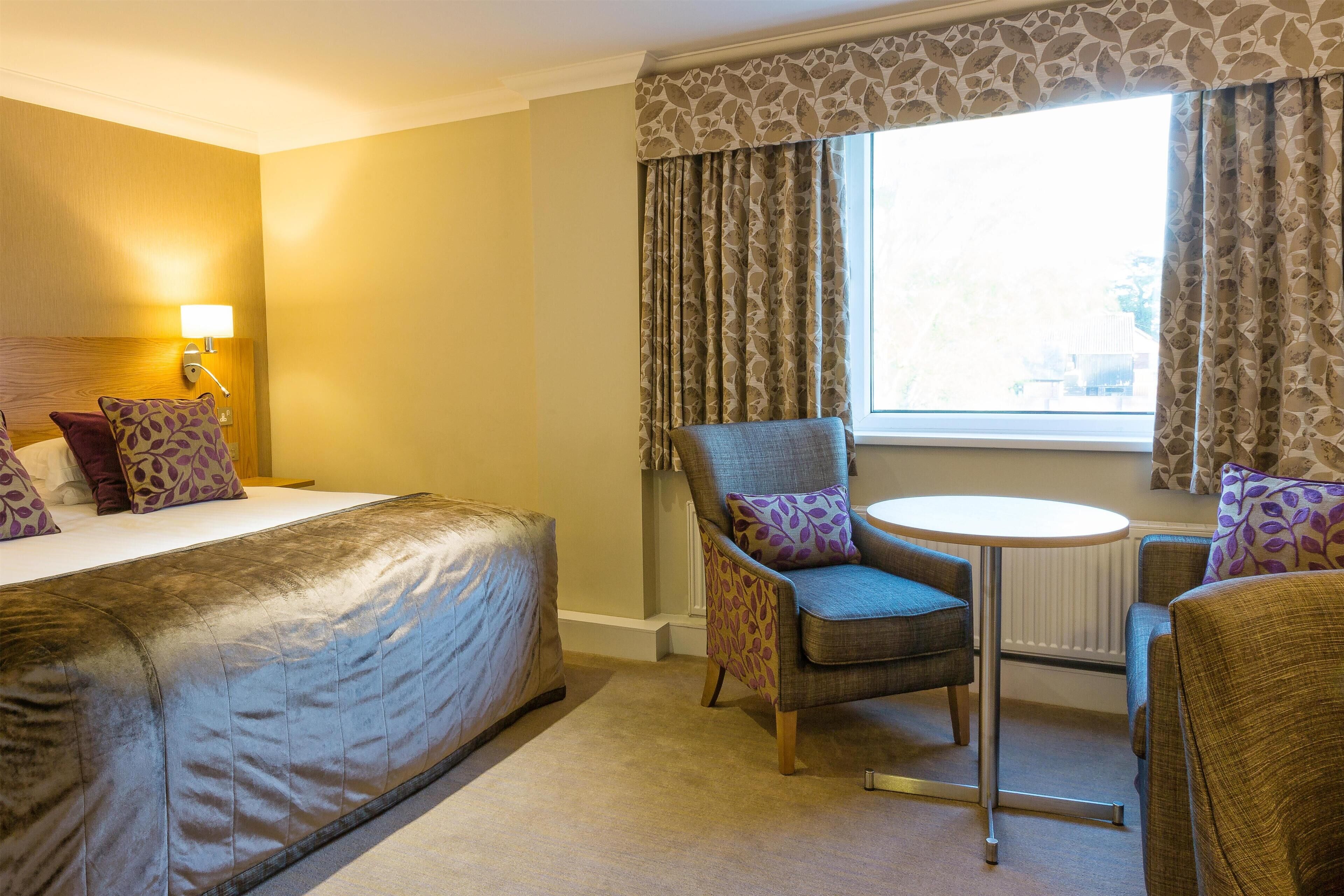 Hotelkamer met een bed, een stoel, een klein tafeltje en een raam met gordijnen.