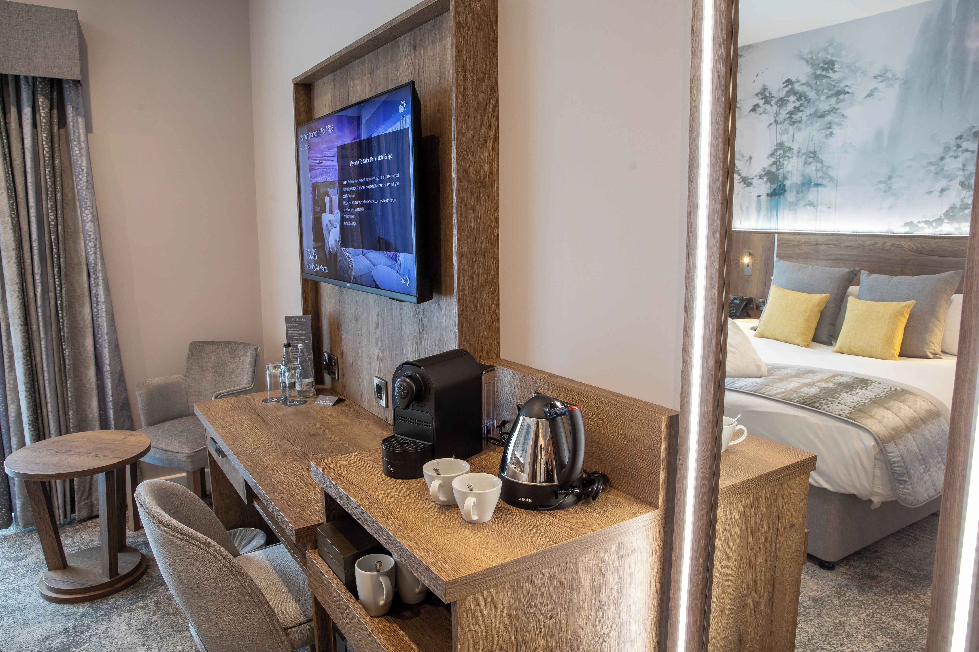 A hotel room with a wooden desk, a coffee maker, two cups, a television mounted on the wall, and a bed with yellow pillows.