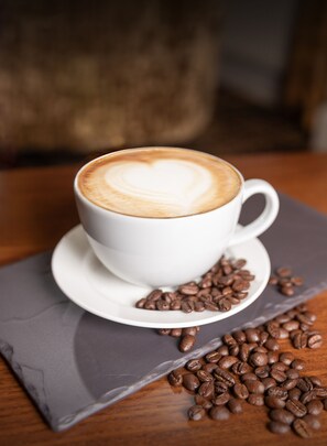 A white cup of coffee with a heart-shaped latte art on a saucer, surrounded by coffee beans on a wooden table.