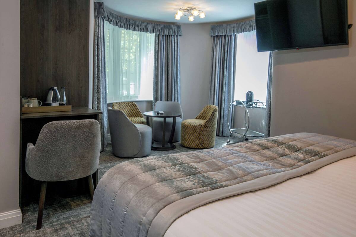 A hotel room with a bed, a desk, two chairs, a small table, and a TV mounted on the wall.