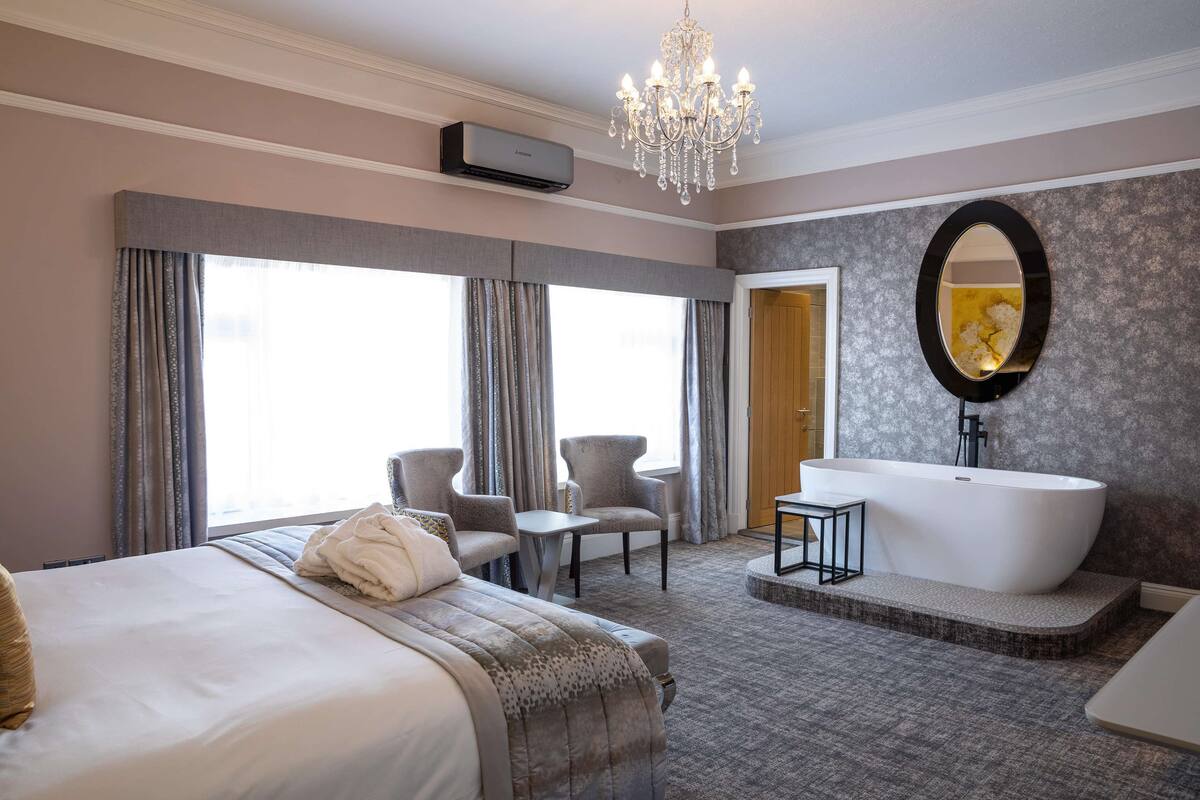 A modern hotel room with a large bed, a sitting area with chairs, a bathtub, and a large mirror.