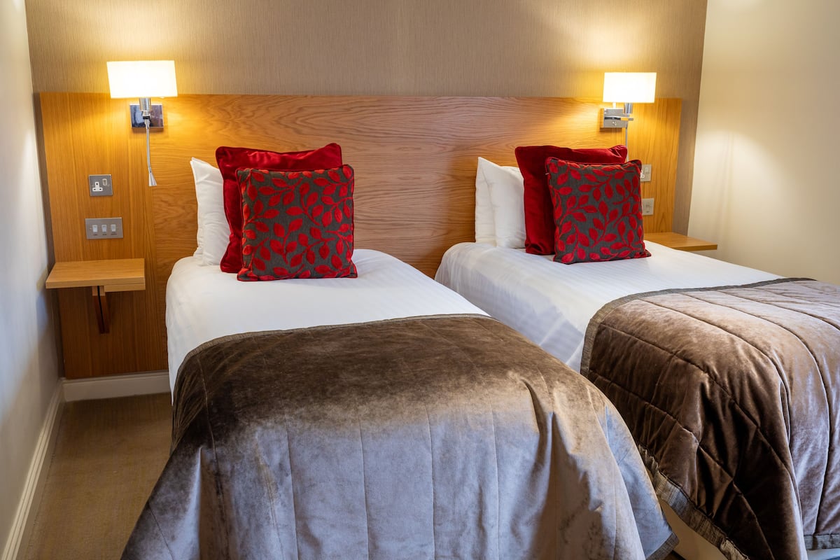 A hotel room with two beds, each with a wooden headboard, white bedding, and red patterned pillows. There are bedside tables with lamps and a power outlet on each side.