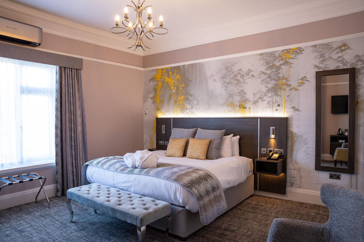A modern hotel room with a large bed, a bench, a bedside table with a lamp, and a wall-mounted TV.