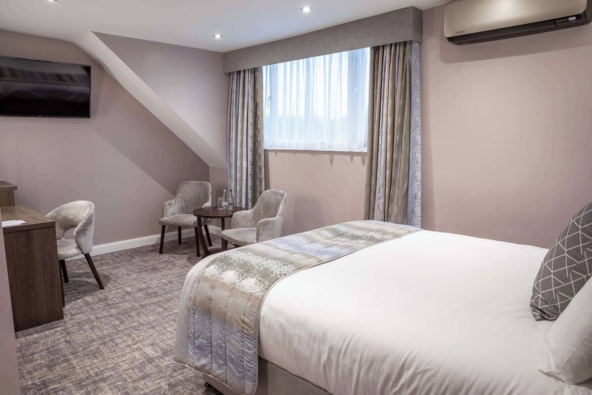 A hotel room with a large bed, two armchairs, a small table, a desk, and a TV mounted on the wall.