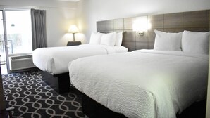 A hotel room with two beds, a patterned carpet, a window with curtains, and a wall-mounted lamp. - Quality Inn Manning I-95 (Manning)