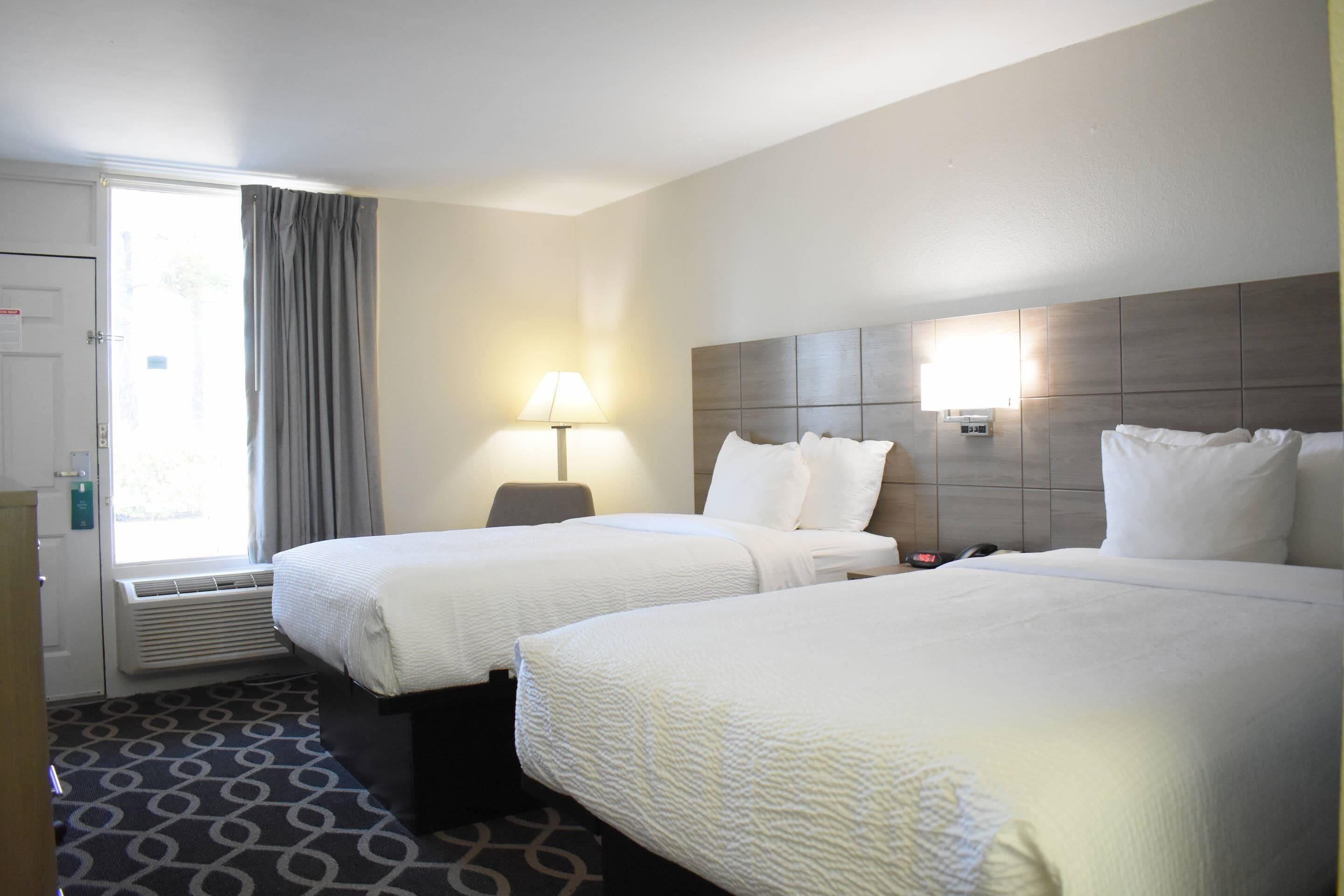 A hotel room with two beds, a window with curtains, a bedside lamp, and a wall-mounted heater.