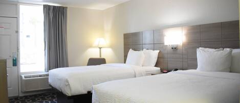 A hotel room with two beds, a window with curtains, a bedside lamp, and a wall-mounted heater.