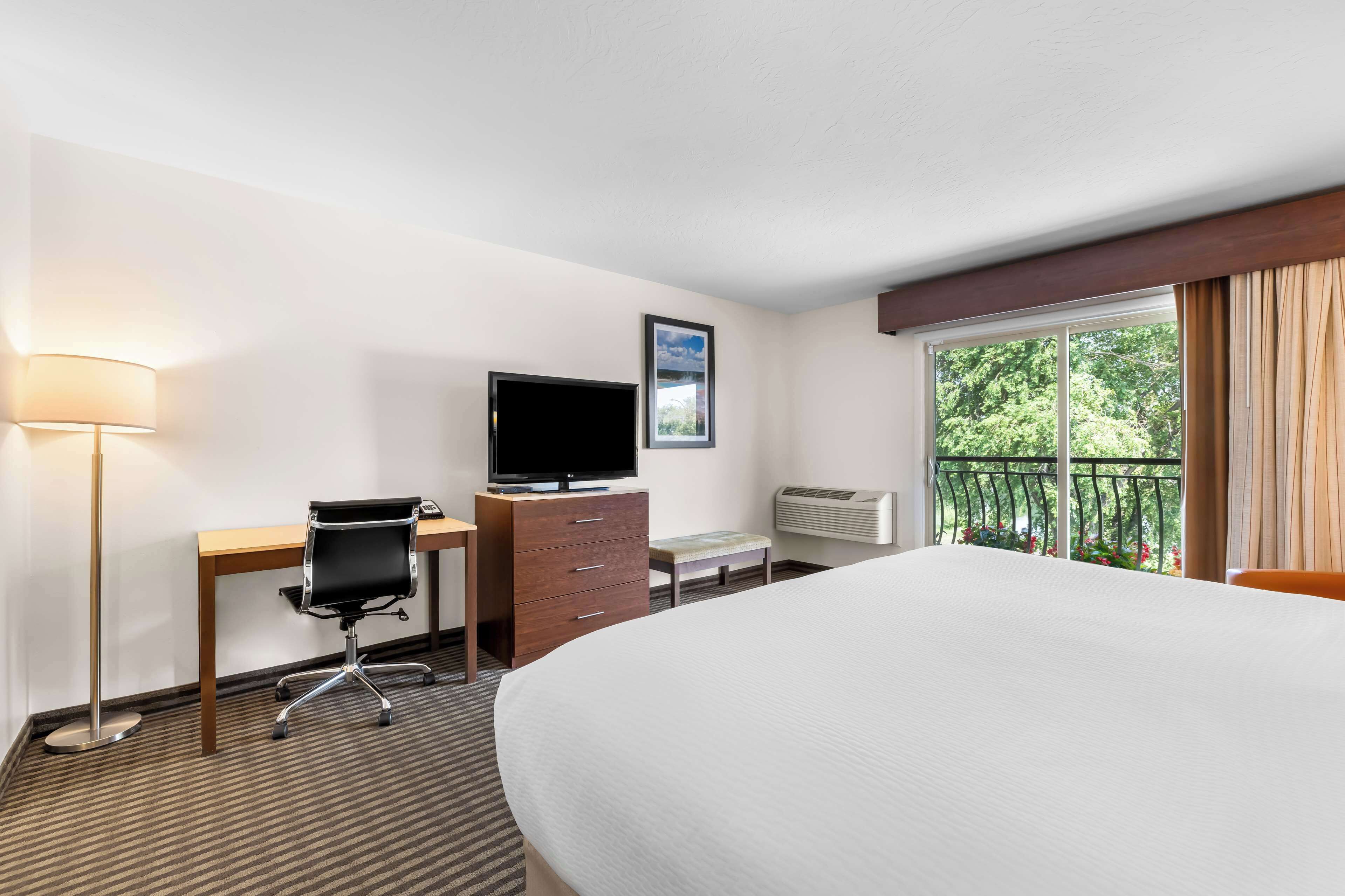 A hotel room with a large bed, a desk with a computer, a TV, a dresser, and a balcony with a view of trees.