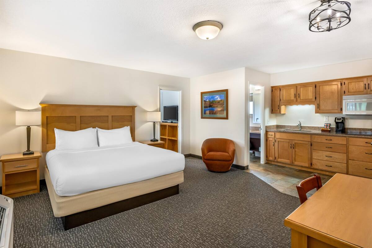 Quarto de hotel com cama, cadeira, escrivaninha, cozinha compacta e banheiro.