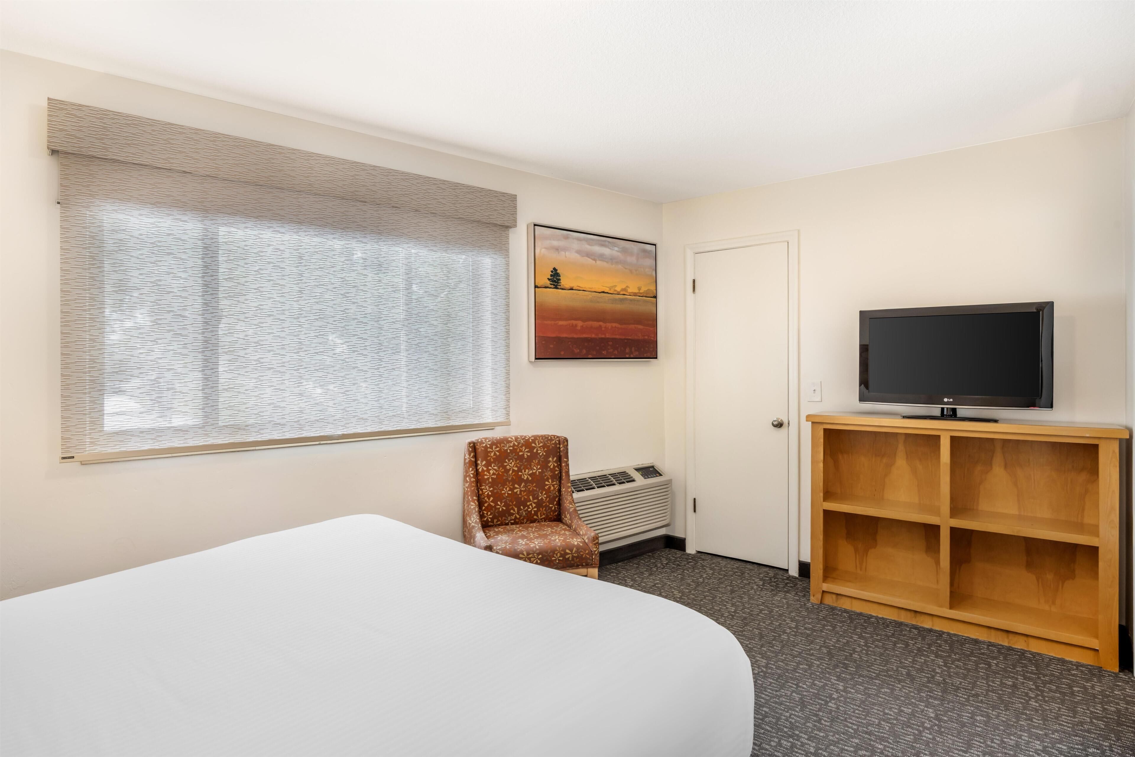 Habitación de hotel con cama, una silla, televisor sobre un soporte de madera, radiador y ventana con persianas.
