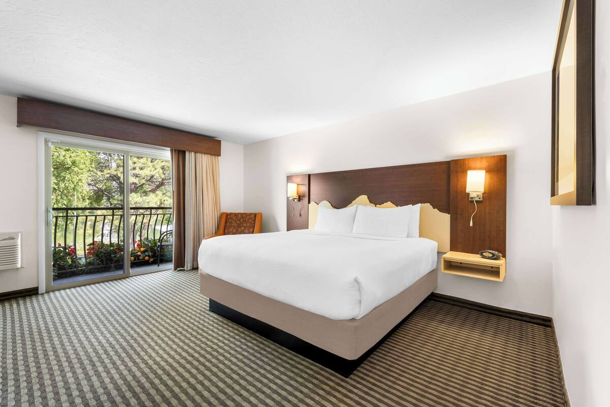 Une chambre d’hôtel avec un grand lit, une table de chevet, une lampe fixée au mur et une porte-fenêtre coulissante donnant sur un balcon agrémenté de plantes.