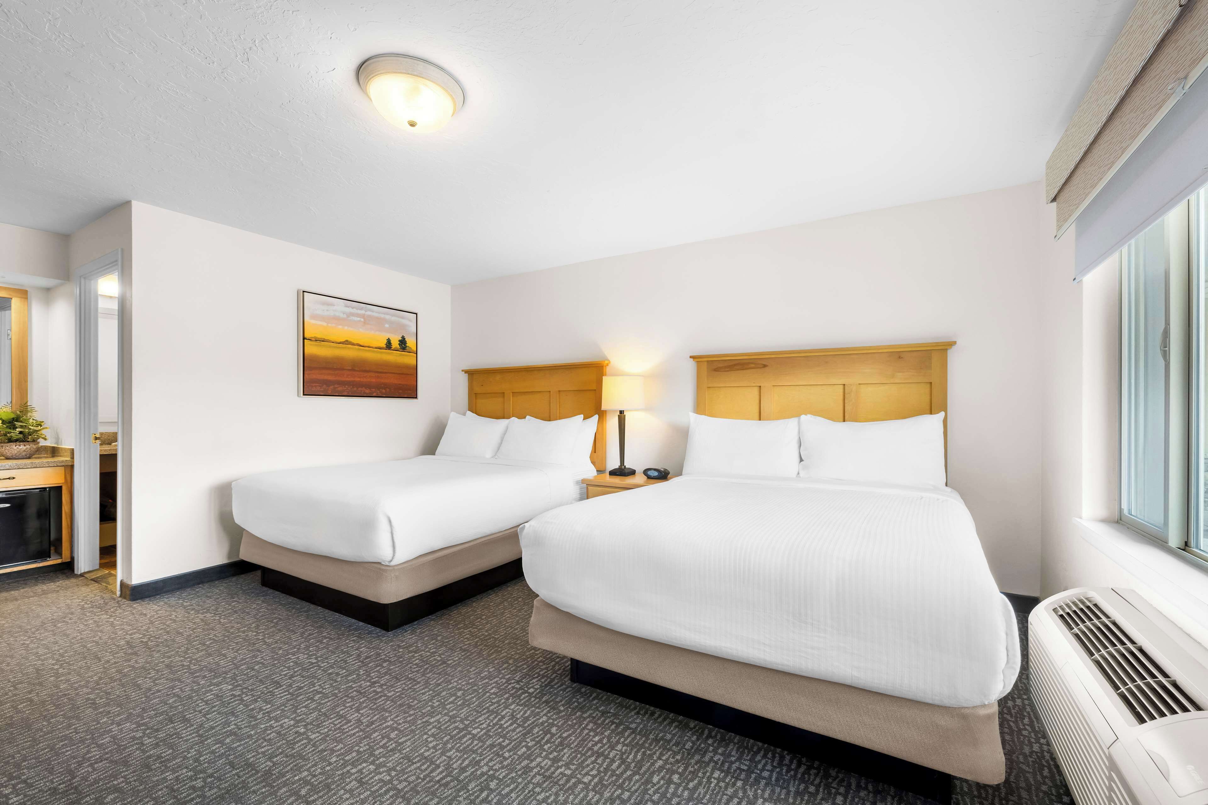 A hotel room with two beds, a bedside table with a lamp, a painting on the wall, and a window with blinds.