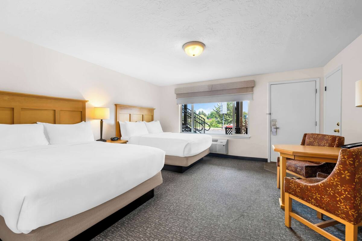 Une chambre d’hôtel avec deux lits, une table à manger avec des chaises, une fenêtre donnant sur un espace verdoyant et une porte.