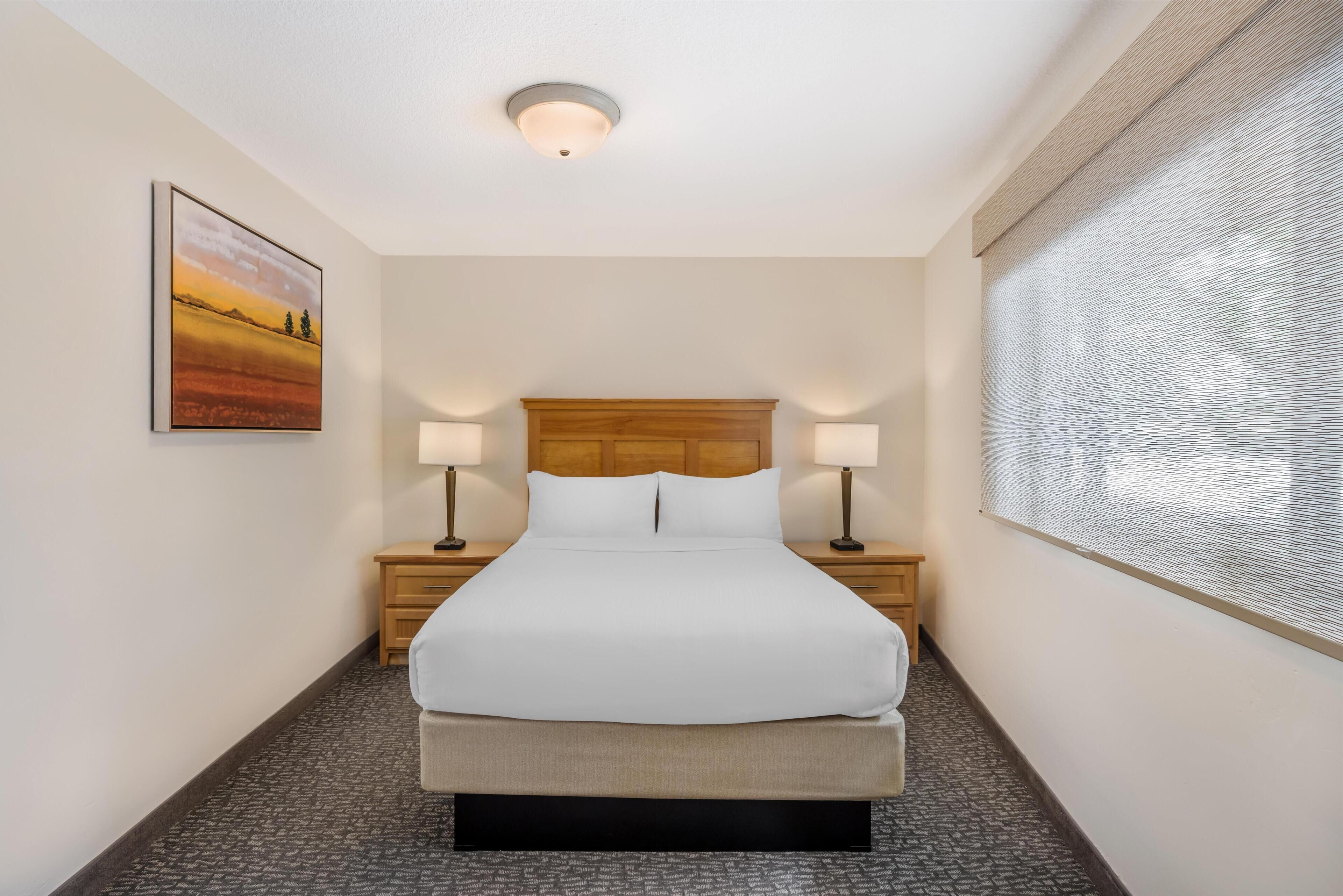 Une chambre d’hôtel avec un lit, deux tables de chevet avec des lampes, une tête de lit, un tableau au mur et une fenêtre avec des stores.