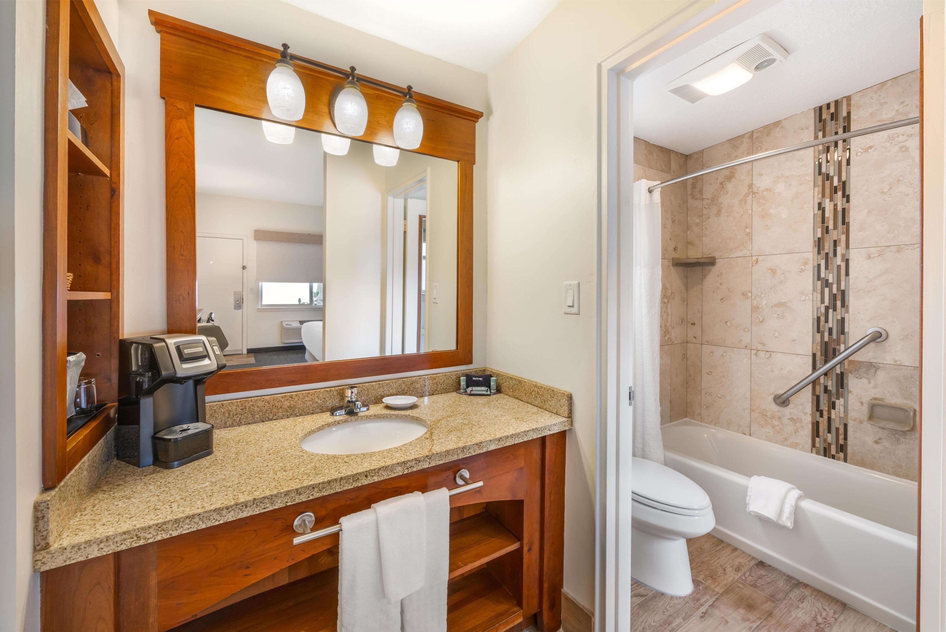 A bathroom with a wooden vanity, a sink, a mirror, a coffee maker, a shower with a glass door, and a toilet.