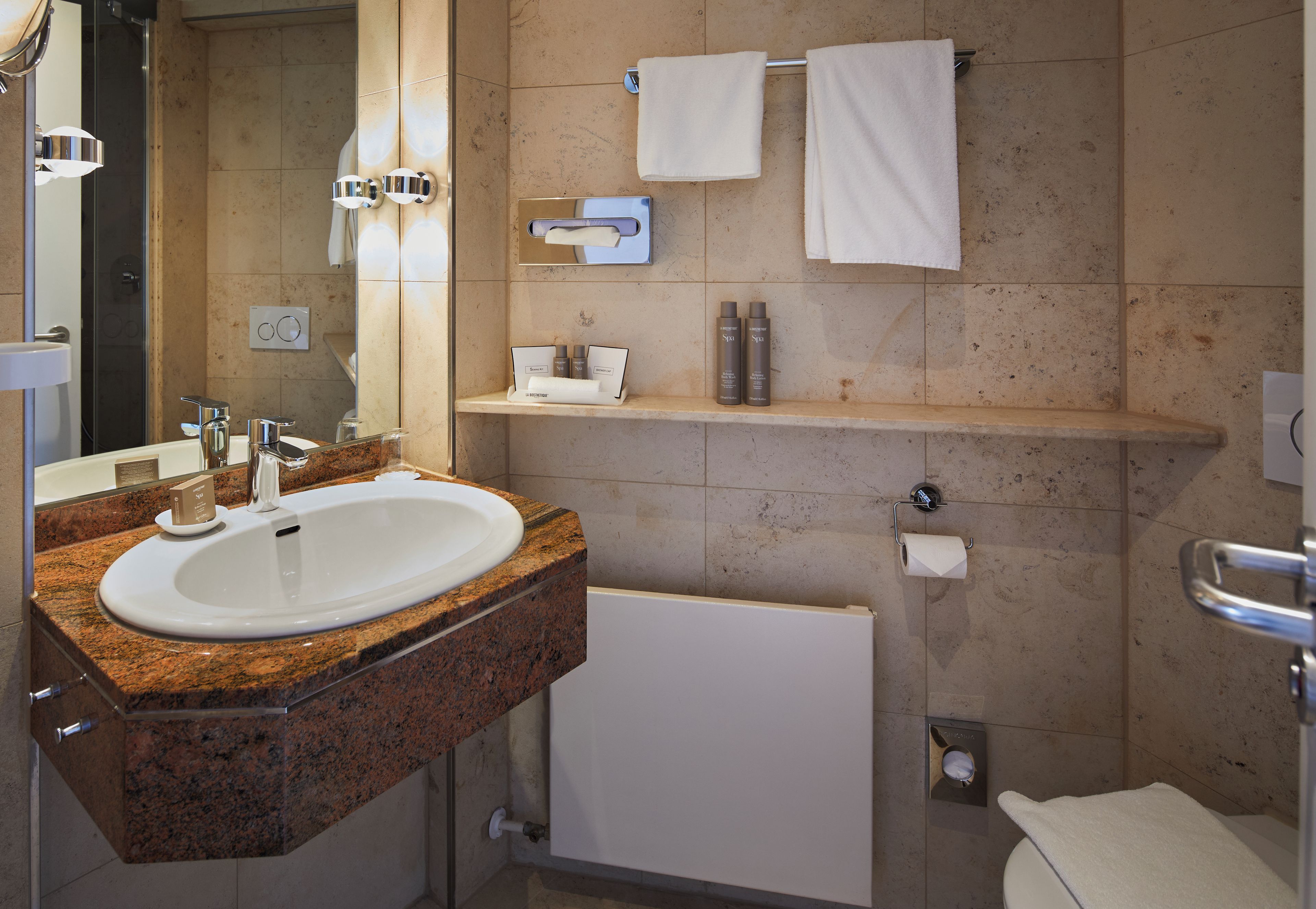 A modern bathroom with a granite countertop sink, a mirror, a towel rack, and a shelf with toiletries.