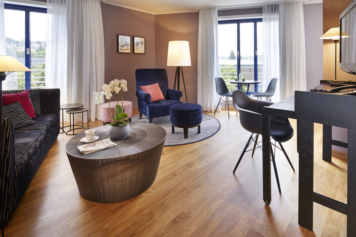 A modern hotel room with a dark blue sofa, a round coffee table, a blue armchair, a dining table with chairs, and large windows with curtains.