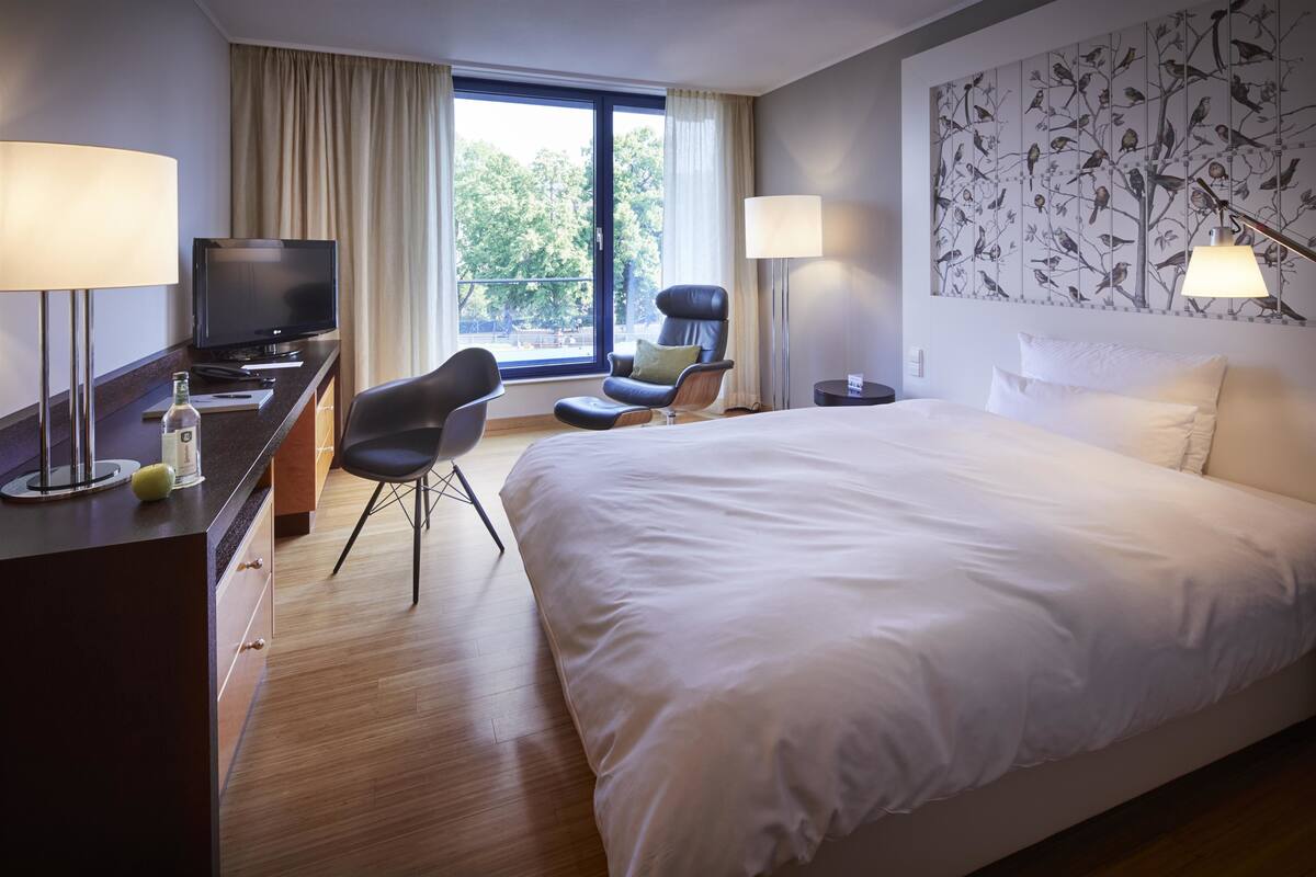 Une chambre d’hôtel avec un grand lit, un bureau avec une télévision, deux chaises et une fenêtre avec des rideaux.