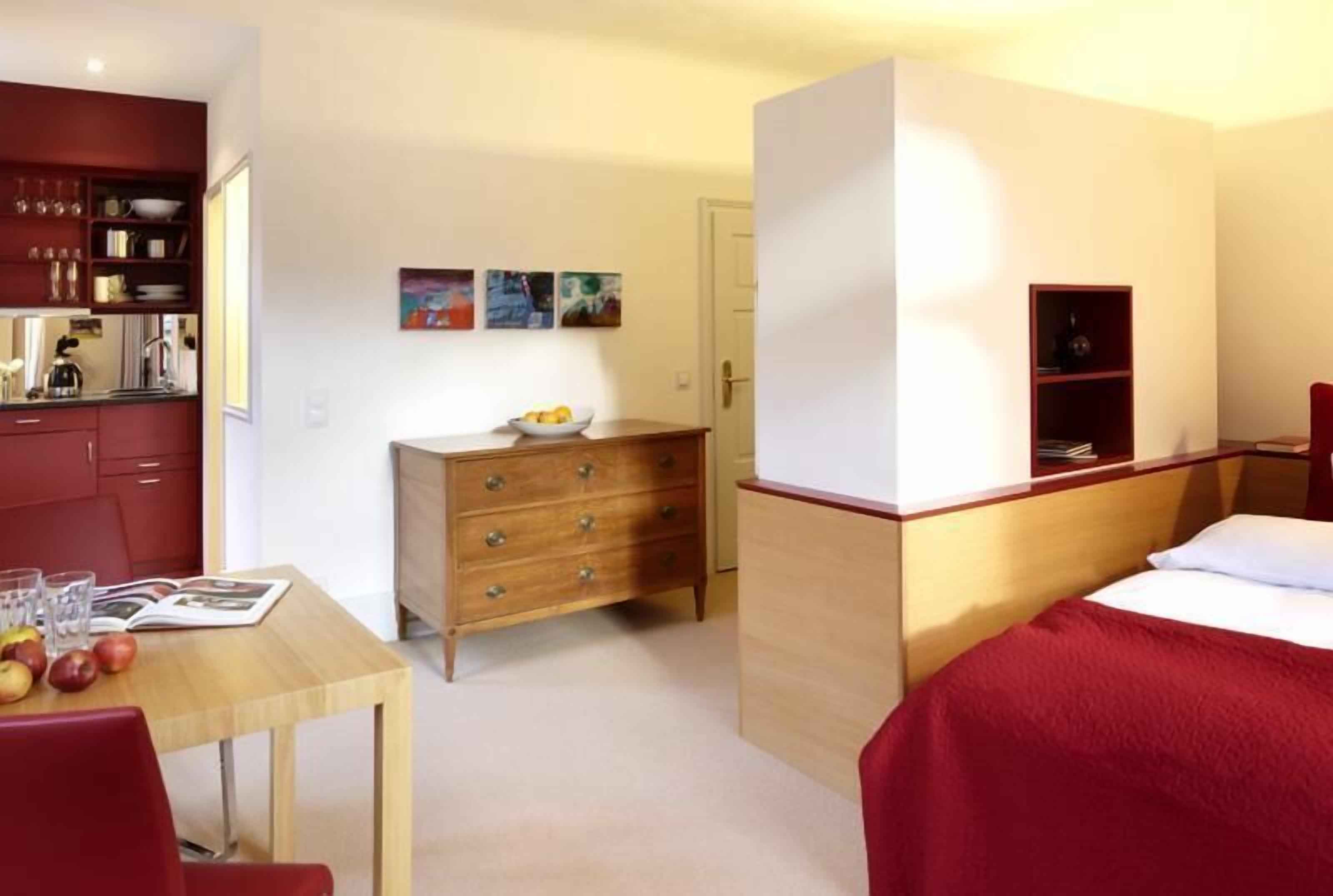 Habitación de hotel pequeña con una cama, un aparador de madera, una mesita con un frutero y una kitchenette.