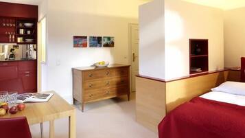 Habitación de hotel pequeña con una cama, un aparador de madera, una mesita con un frutero y una kitchenette.