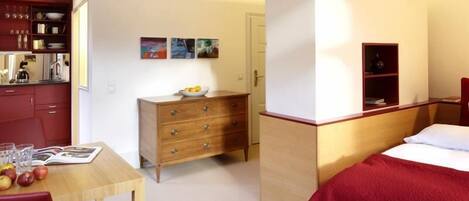Habitación de hotel pequeña con una cama, un aparador de madera, una mesita con un frutero y una kitchenette.