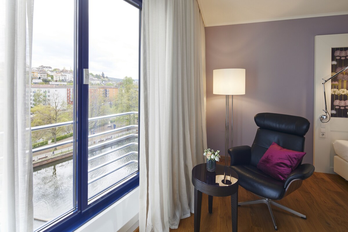 A modern room with a view of a cityscape through a large window, a black leather chair with a purple pillow, a small side table with a vase, and a floor lamp.