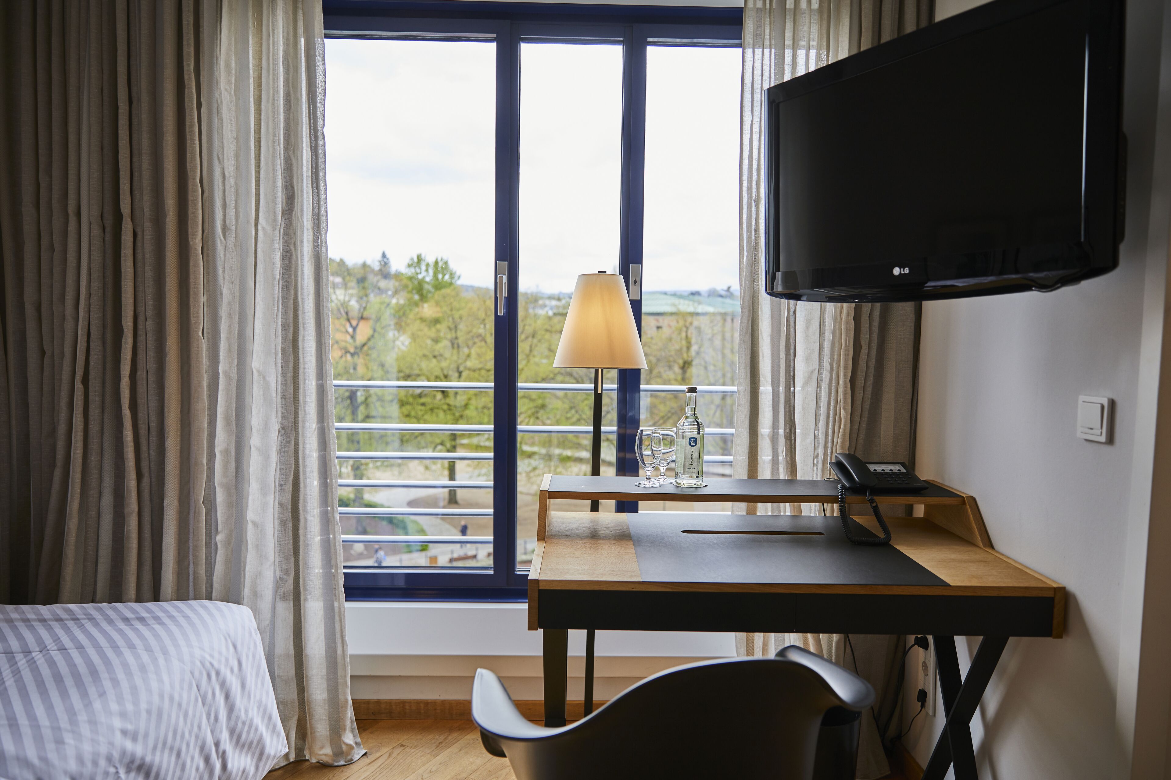 A hotel room with a bed, a desk with a lamp, a telephone, and a TV.