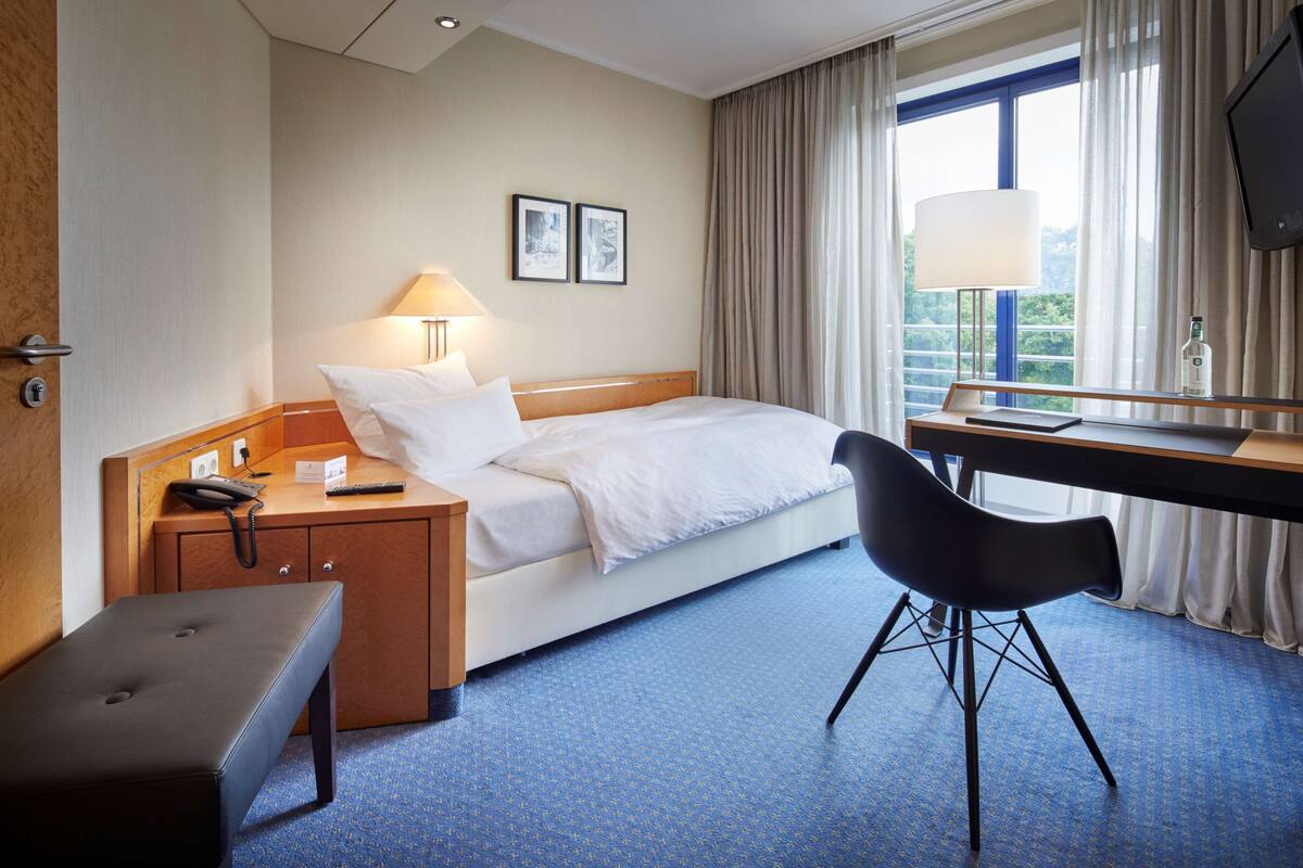 Une chambre d’hôtel comprenant un lit, une table de chevet avec une lampe, un bureau avec une chaise, un téléphone et une fenêtre avec des rideaux.